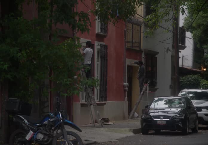 Two men on ladders renovate the exterior of a colorful residential building on a tree-lined city street