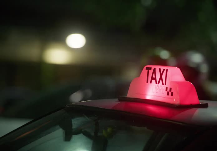 A red illuminated taxi sign glows on a car roof amidst the bustling, blurry lights of city traffic at night