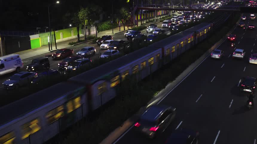 A brightly lit passenger train travels alongside a congested highway full of cars during the evening rush hour