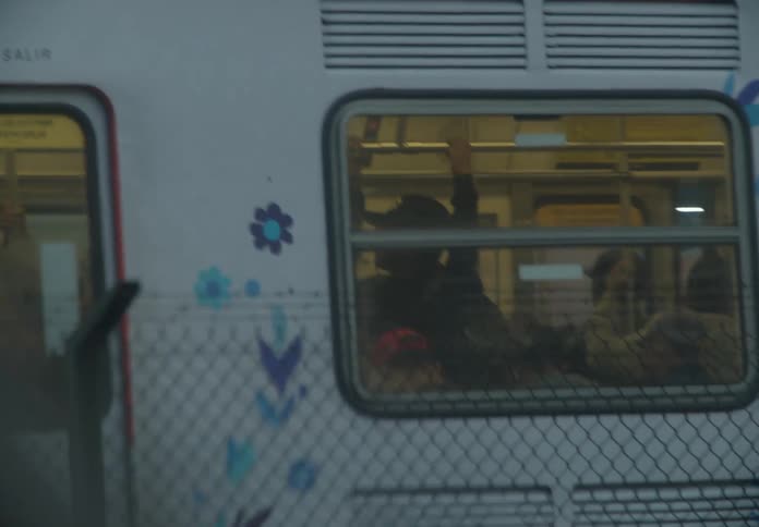 A colorful subway train filled with commuters moves past the camera, viewed through a chain-link fence