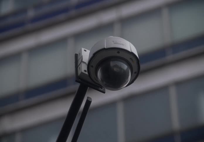 A slow zoom-in on a dome security camera mounted on a pole with a modern office building in the background