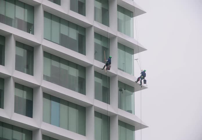 Two window washers rappel down the side of a modern white office building to clean the glass facade