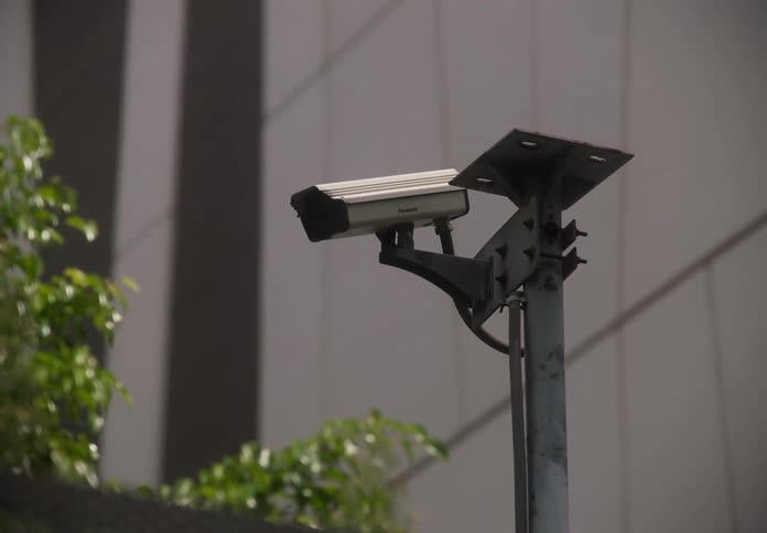 A security camera on a pole slowly zooms out to show an urban setting with a modern building and fence