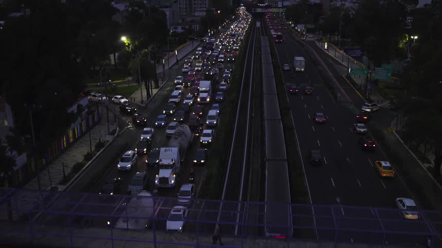Ascending aerial view of a major city highway at night shows heavy rush hour traffic, a pedestrian overpass, and a moving train