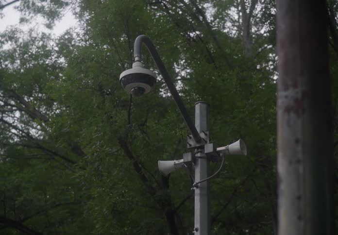 A surveillance camera and loudspeakers on a pole stand against a backdrop of trees swaying on a windy, overcast day