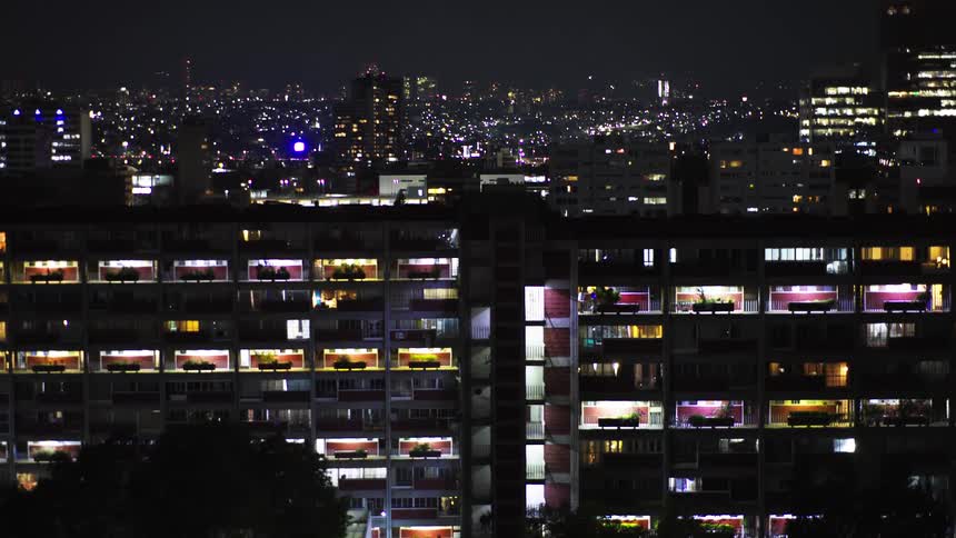 A time-lapse view of a brightly lit apartment building at night, with a slow zoom-out revealing the expansive, glowing city skyline behind it