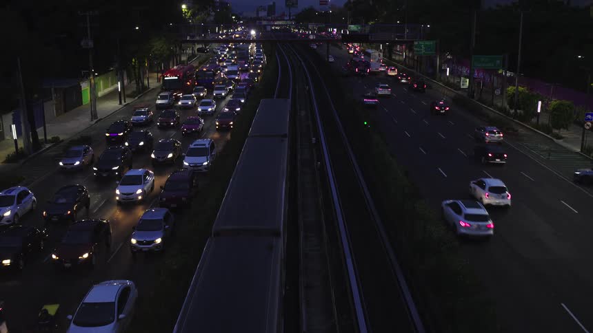 A high-angle view shows a stopped train on the tracks while heavy rush hour traffic with headlights on moves slowly along a major city highway at dusk