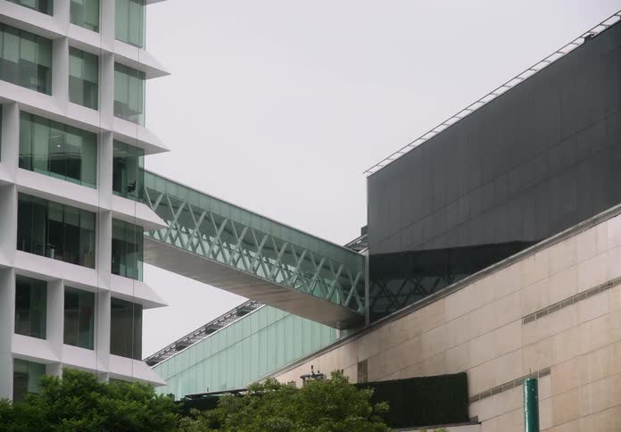 A slow zoom focuses on a modern glass skywalk connecting two contemporary office buildings in a bustling city on an overcast day