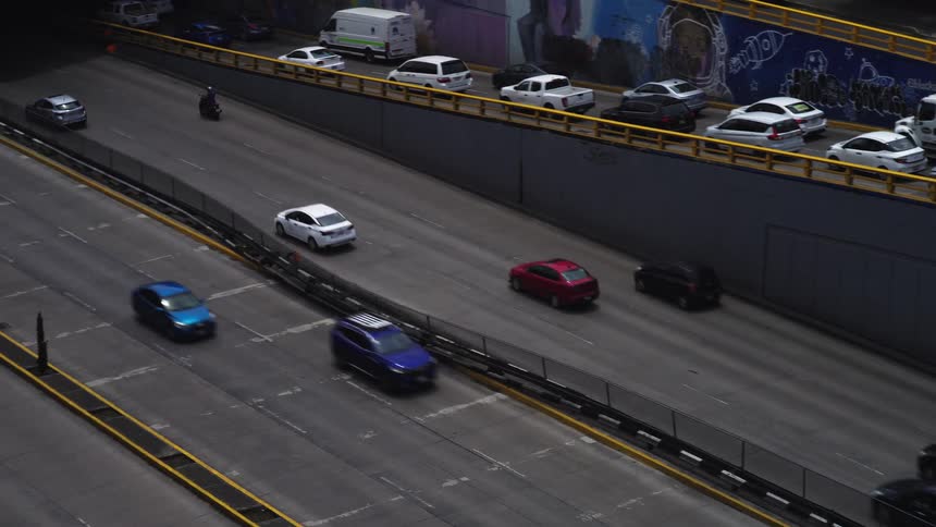 A high-angle, slow zoom-out shot of a multi-level urban freeway showing cars and motorcycles flowing into a tunnel
