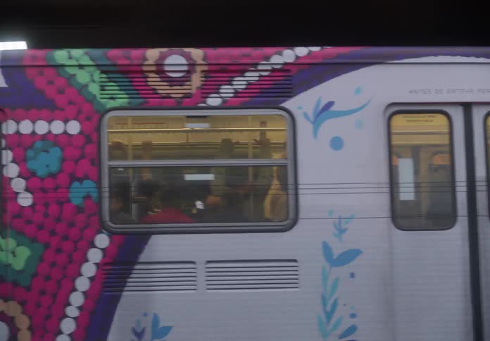 A tracking shot of a decorated Mexico City Metro train passing by, revealing passengers commuting inside