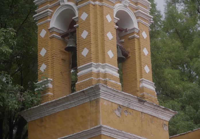 A slow tilt up reveals a historic yellow church bell tower with a cross on top, nestled among trees on a misty day