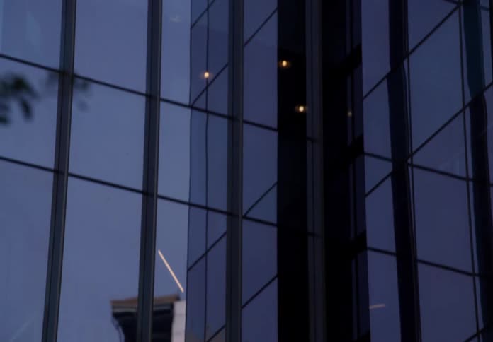 A slow zoom-in on the reflective glass facade of a modern skyscraper under an overcast sky, revealing other city buildings in the reflection