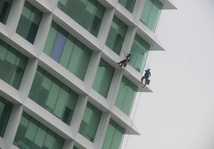 Two professional window washers rappel down a modern skyscraper to clean its glass facade on an overcast day