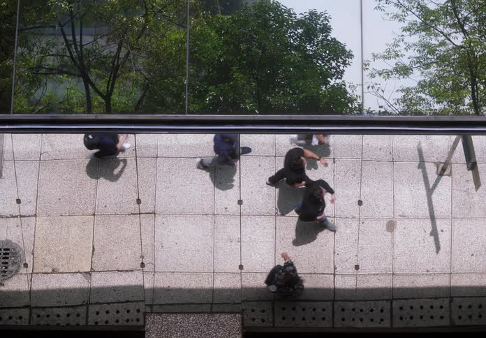 A high-angle, top-down view of various pedestrians walking along a city sidewalk casting shadows