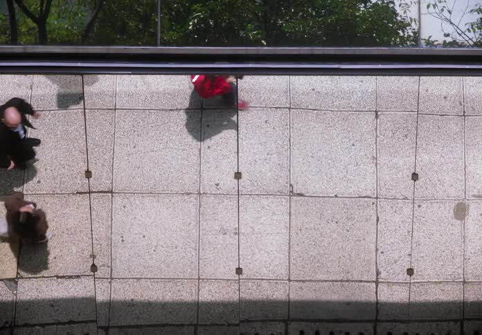 A high-angle tilt reveals a bird's-eye view of anonymous city dwellers walking on a sidewalk below a reflective building