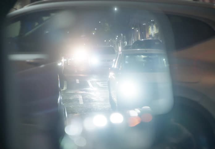 A dreamy point-of-view shot looking into a car's side mirror reveals the bright headlights of nighttime city traffic