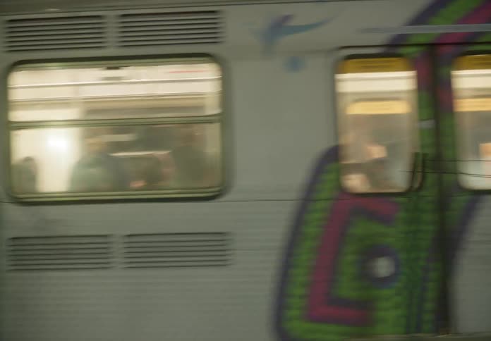 Tracking shot of a graffiti-covered subway train passing through a station at night, with passengers seen through the windows
