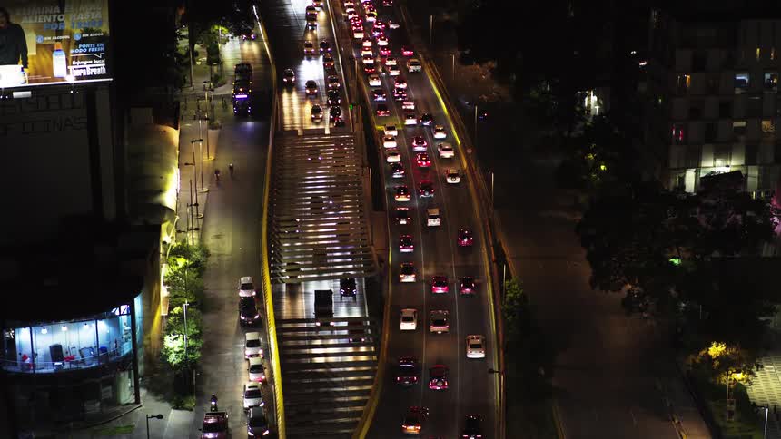 An elevated view shows a bustling city avenue at night, with slow-moving traffic and pedestrians on an overhead bridge