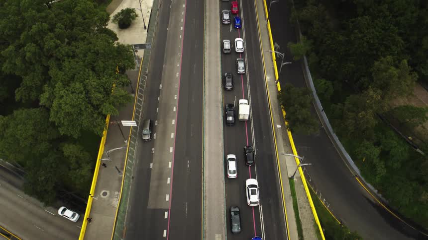 An aerial top-down view shows slow-moving traffic on a city highway overpass surrounded by lush green trees
