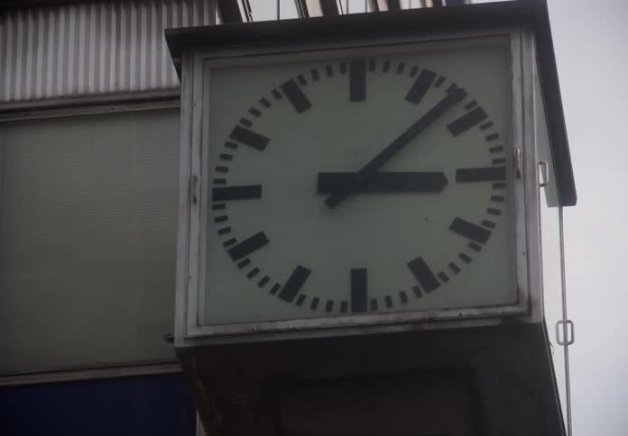 A slow zoom in on a large, square clock mounted on the side of an urban building