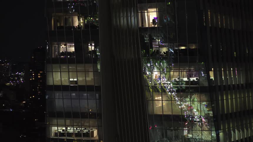 Aerial view zooms out from an empty office inside a modern glass skyscraper to reveal the sprawling city lights and traffic at night