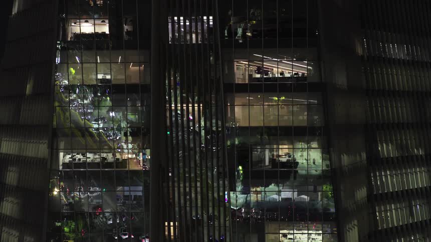 An aerial shot reveals the exterior of a modern office tower at night, with the bustling city life reflected in its windows