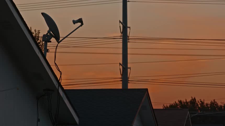 A satellite dish sits on a rooftop in a suburban neighborhood, silhouetted against the warm orange sky at dusk