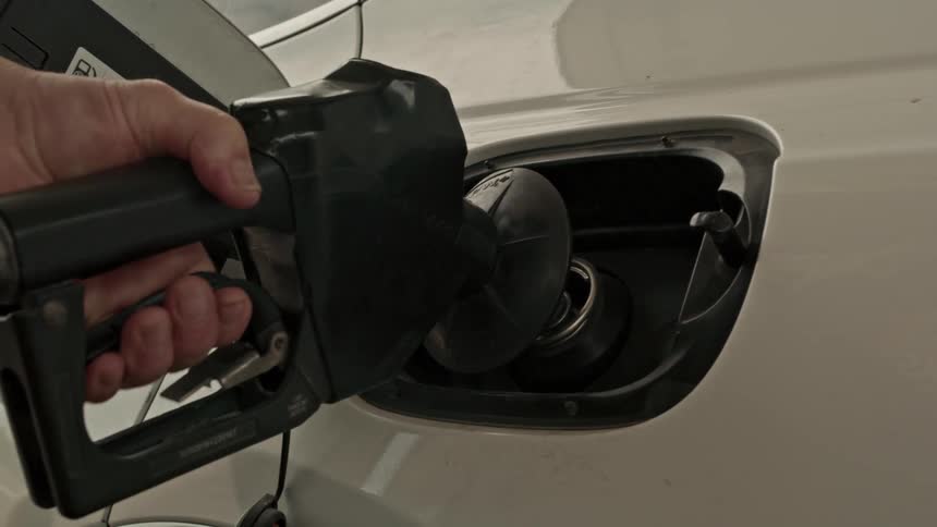 A person's hand opens the fuel cap of a white car and inserts the pump nozzle to refuel with gasoline