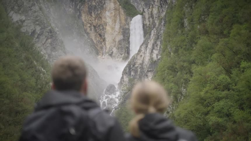 An out-of-focus couple in jackets observes a powerful waterfall cascading down a rocky cliff into a lush, green canyon