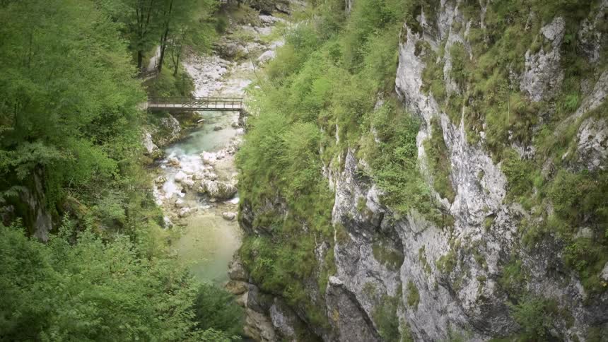 A serene, wide shot of a wooden footbridge crossing a clear river at the bottom of a lush, rocky canyon