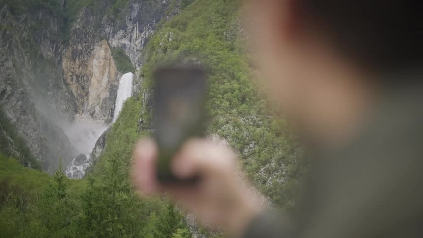 A person records a stunning mountain waterfall on their smartphone with a smooth focus pull from the device to the beautiful landscape