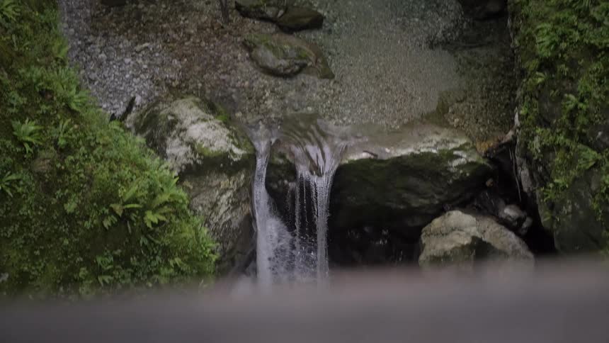 A high-angle slow reveal of a hidden waterfall cascading down mossy rocks into a clear stream in a lush green gorge