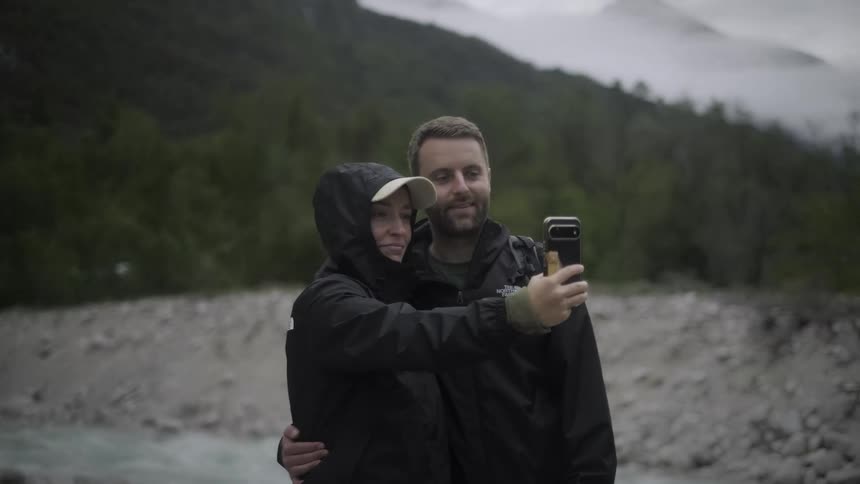 A happy couple in rain jackets takes a selfie together in a scenic mountain valley near a river