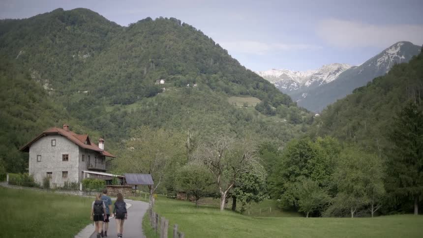 A trio of hikers follows a path through a lush green valley, passing a rustic stone house with towering mountains in the background