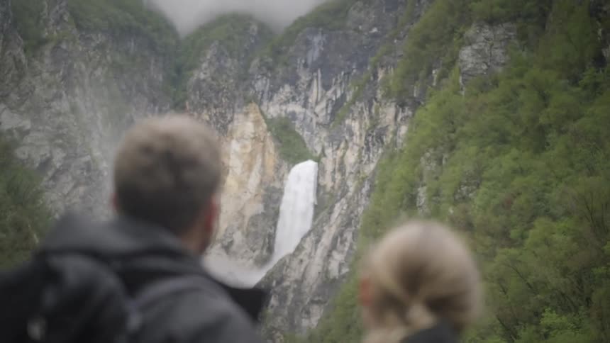A smiling couple in hiking gear turns in slow motion to admire a powerful waterfall in a lush mountain landscape