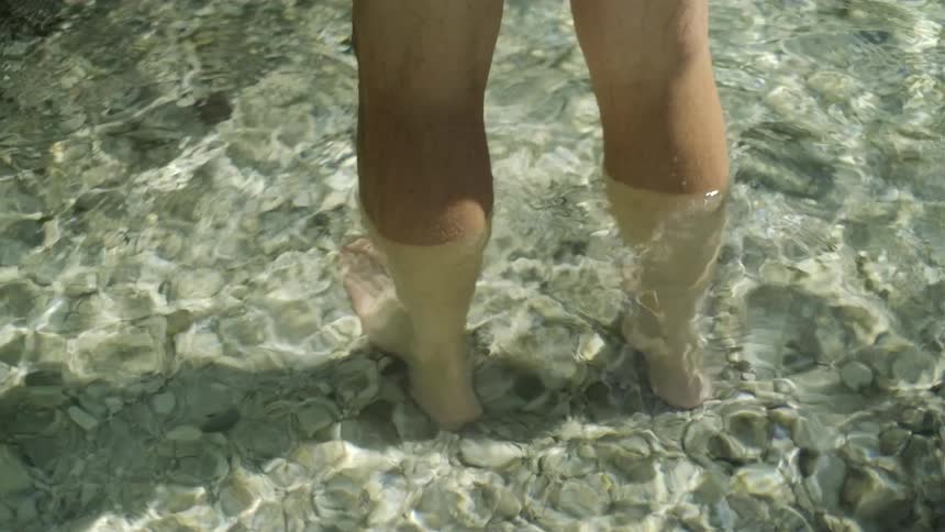 A person's bare feet and legs stand in the clear, shallow water of a rocky riverbed as sunlight shimmers on the surface