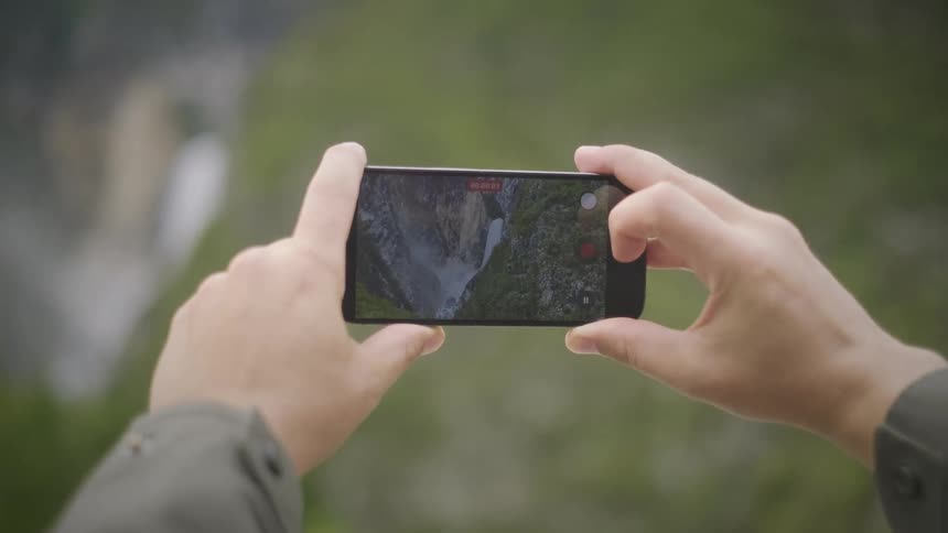 A person's hands hold a smartphone, pulling focus to record a beautiful waterfall cascading down a mountain in a lush green valley