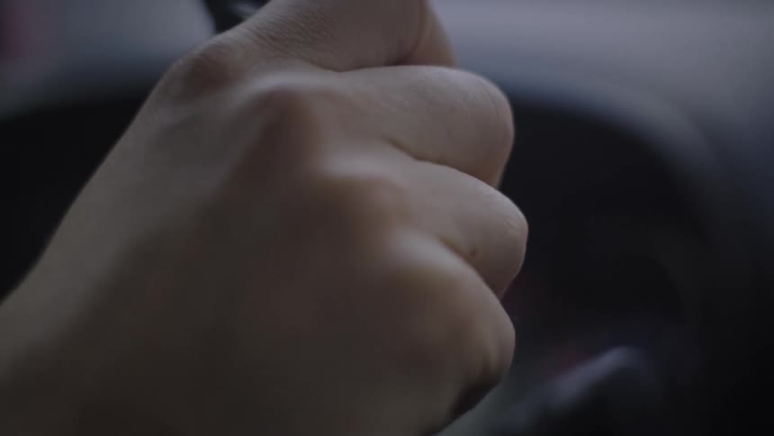 A somber close-up shot of a hand gripping a car's steering wheel in low light