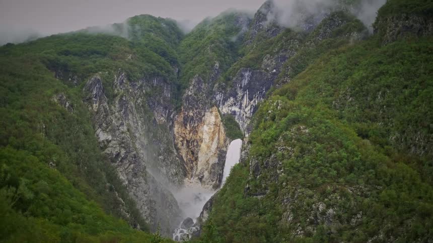 A powerful waterfall cascades down a rocky cliff face in a lush green mountain valley shrouded in mist