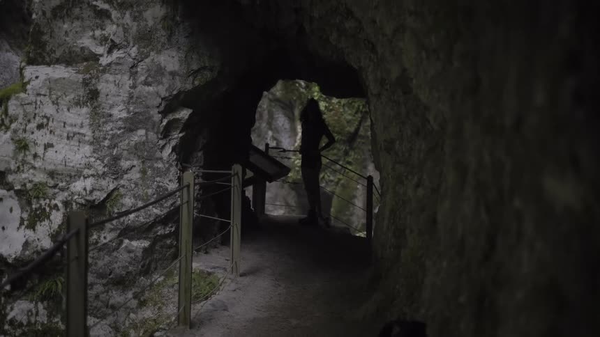 A silhouetted person stands in a dark, rocky cave tunnel, looking out towards the light at the end