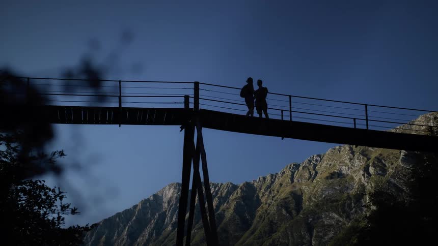 A silhouetted couple shares a romantic moment on a high suspension bridge overlooking a scenic mountain range