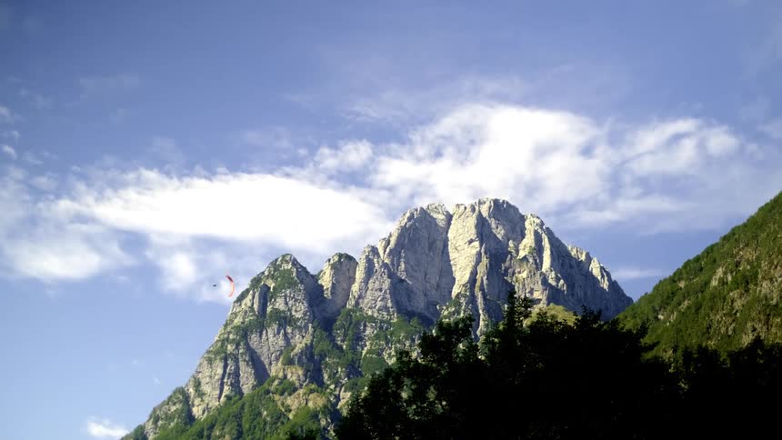 A red paraglider soars peacefully across a blue sky in front of a majestic, tree-lined mountain range