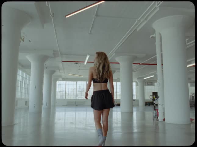 A stylish woman in workout attire walks through a large, empty industrial loft and turns to look back over her shoulder