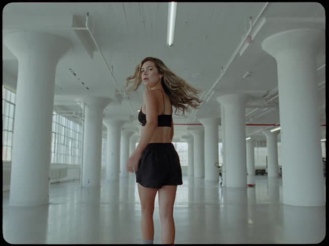 Slow motion shot from behind of a fit woman walking away through a large, empty industrial loft before turning to look back at the camera