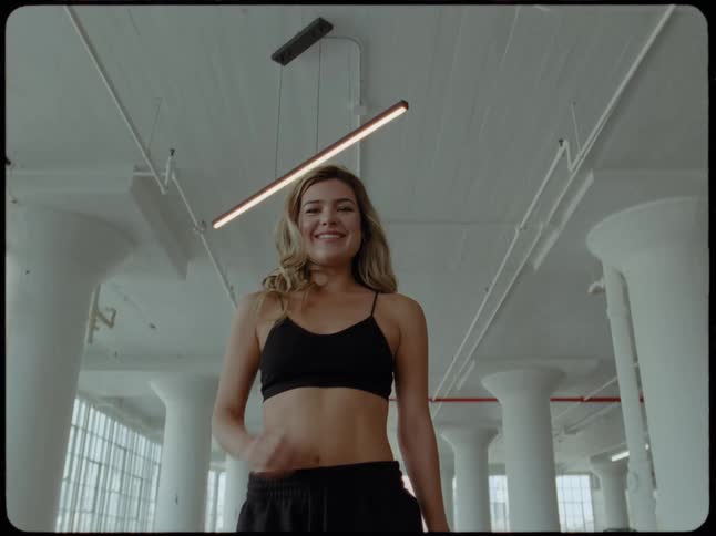 A stylish woman in workout attire walks towards the camera and smiles before turning away in a bright, industrial loft