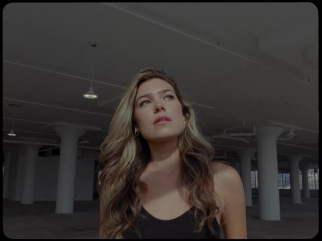 A beautiful woman with long wavy hair stands thoughtfully in a large, empty industrial loft, her face illuminated by a soft light