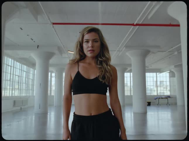 A stylish woman in activewear confidently walks toward the camera in a large, empty industrial studio loft