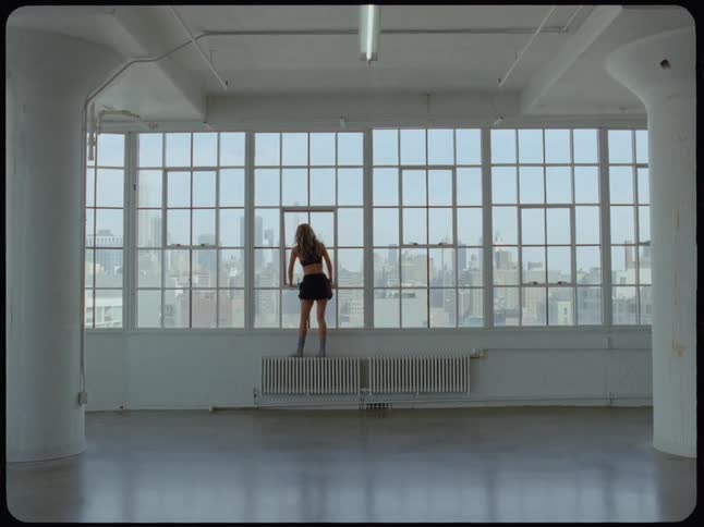 Wide shot slowly zooms in on a woman standing on a radiator, looking out a large window at the city skyline from an empty industrial loft