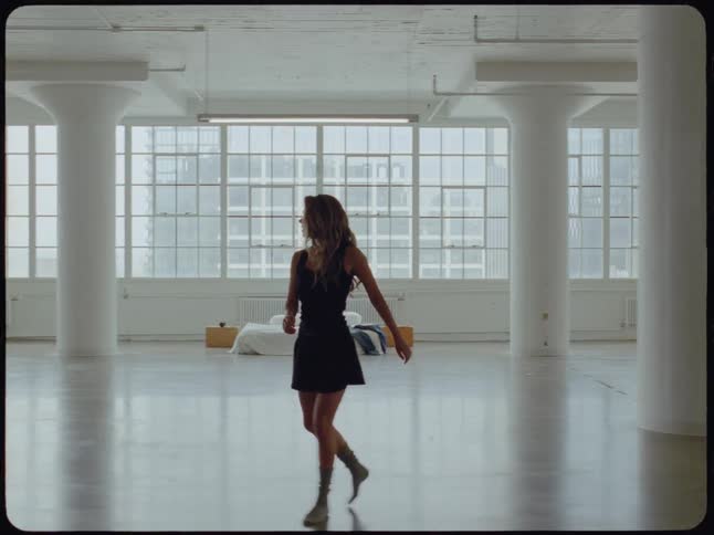 A solitary woman walks and dances thoughtfully through a vast, sunlit, minimalist studio space with large industrial windows