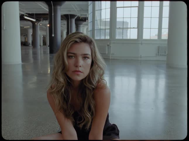 A beautiful and pensive woman with long hair sits on the concrete floor of an empty loft and looks at the camera